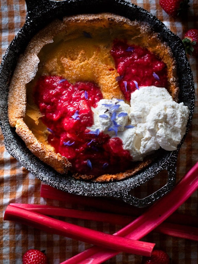 picture of a dutch baby in a cast iron skillet on a checkered tablecloth with jam and whipped cream